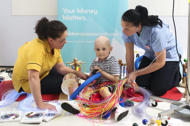Two women engage with a child playing with colorful tubes and toys, in front of a banner that reads "Your Money Matters" and "Children's Cancer Unit." The new arrival of multisensory toy boxes adds a vibrant touch to the Children's Cancer and Haematology Ward.