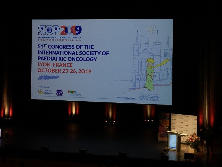 Stage with a large screen displaying "51st Congress of the International Society of Paediatric Oncology" in Lyon, France, October 23-26, 2019. The lectern adorned with flowers sets the scene as Dr. Lyndsey Thompson highlights key moments from SIOP 2019.