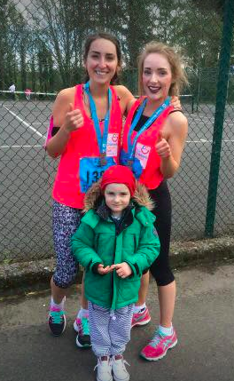 Two women wearing race medals pose happily with a child in a green coat, proudly standing before a chain-link fence near a sports area after completing their New Year's resolution to raise funds for charity.