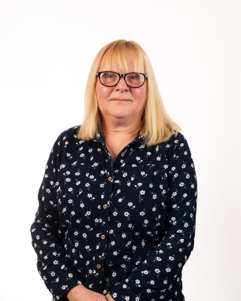 A person with shoulder-length blonde hair, wearing glasses and a dark, flower-patterned button-up shirt, stands against a plain white background, exuding an air of leadership.