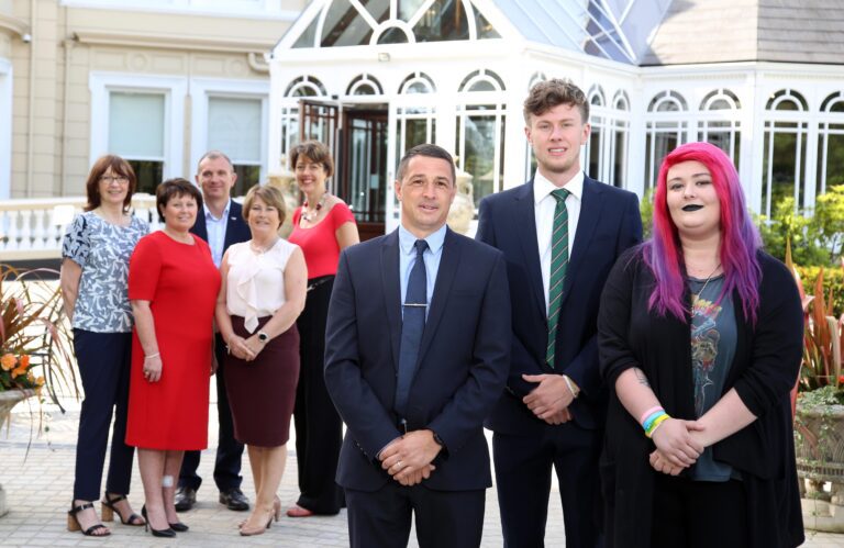 A group of young adults stands outside a building with large windows. Two men in suits stand in front, while others in business attire are in the background.
