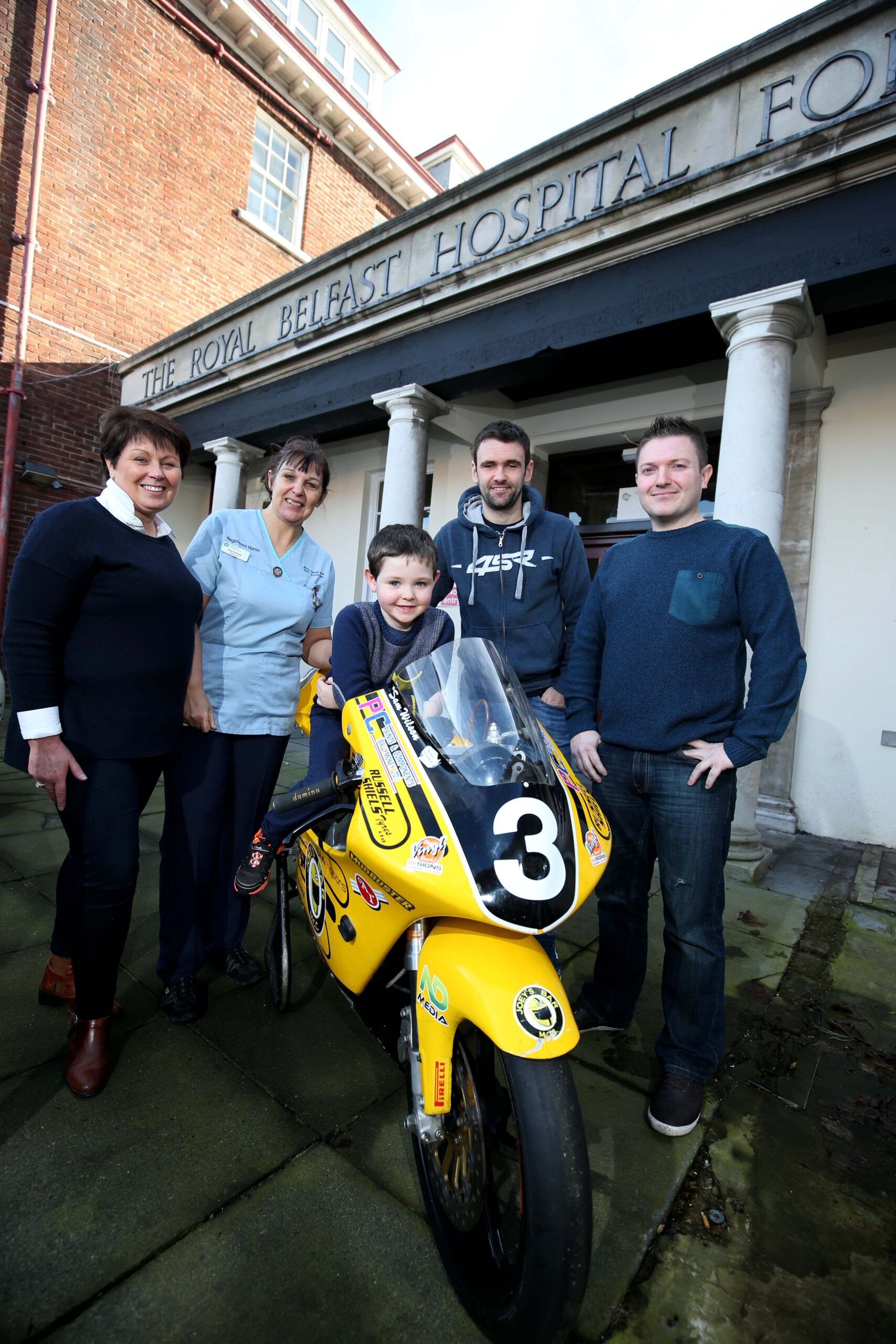 A group of four adults and one child proudly pose with a small yellow motorcycle in front of a building labeled "The Royal Belfast Hospital," symbolizing their shared goals to support the Children's Cancer Unit.