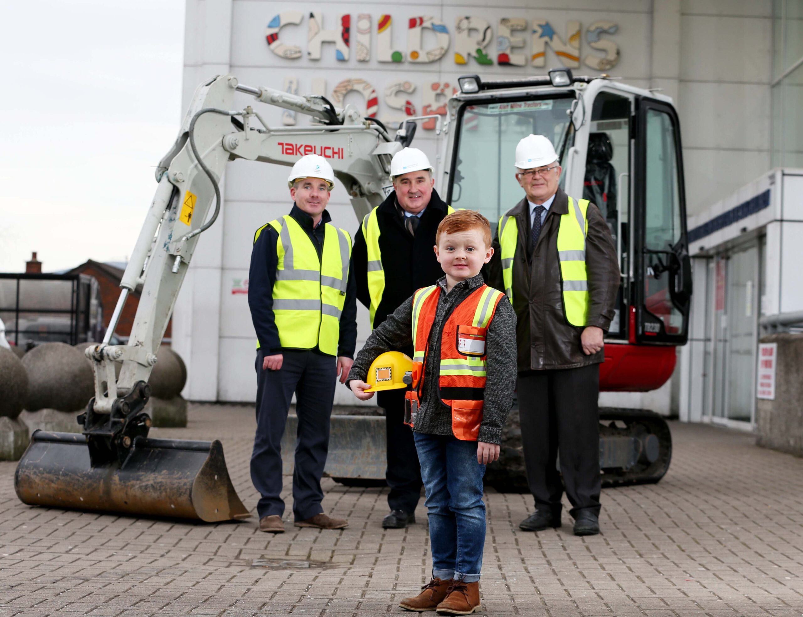 A child in a construction vest and hard hat stands in front of a mini excavator and three adults in safety gear outside the RBHSC building with "Children's" on the sign, as part of the Children’s Cancer Unit Charity initiative to build new bedrooms.