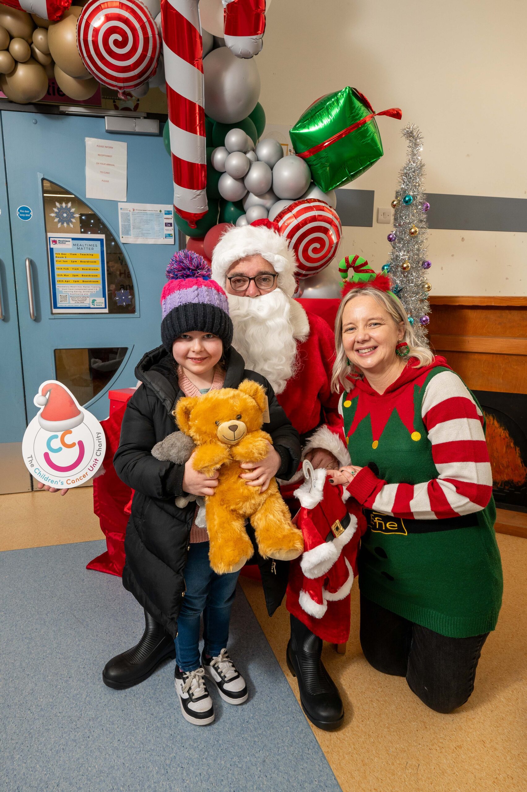 A child holding a teddy bear, part of Zoe and Sophie's story, poses with a person dressed as Santa and another in an elf costume, standing next to holiday decorations indoors. This heartwarming scene raises awareness for International Childhood Cancer Day.