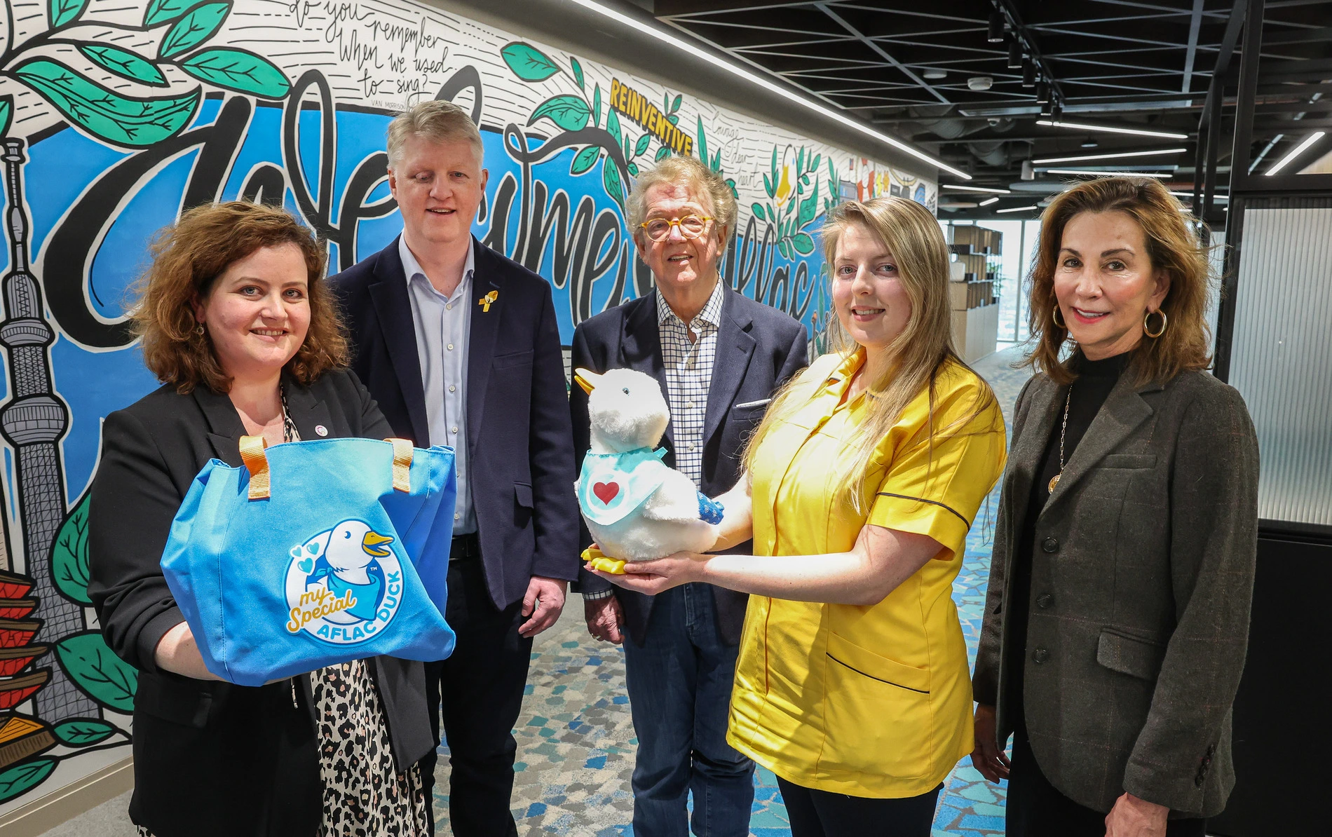 A group of five people posing indoors, showcasing their creative partnerships. One holds a plush duck toy, another a tote bag, with a colorful wall mural in the background.