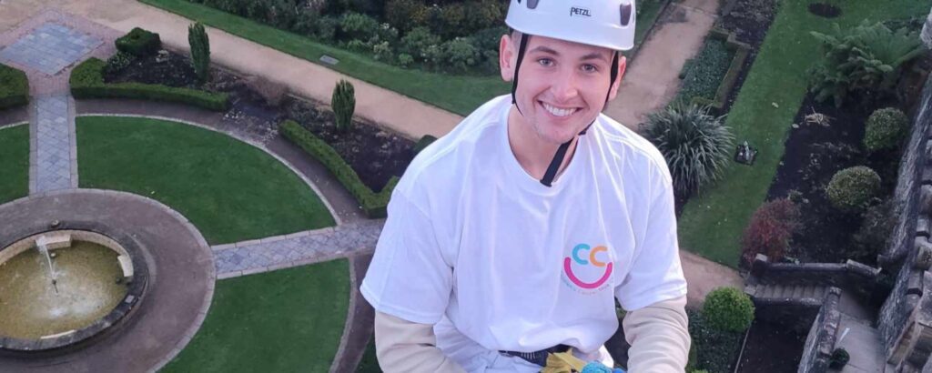 A person in a white helmet and t-shirt participates in an exhilarating Europa Abseil adventure activity, gracefully descending down a building with a lush garden visible below.