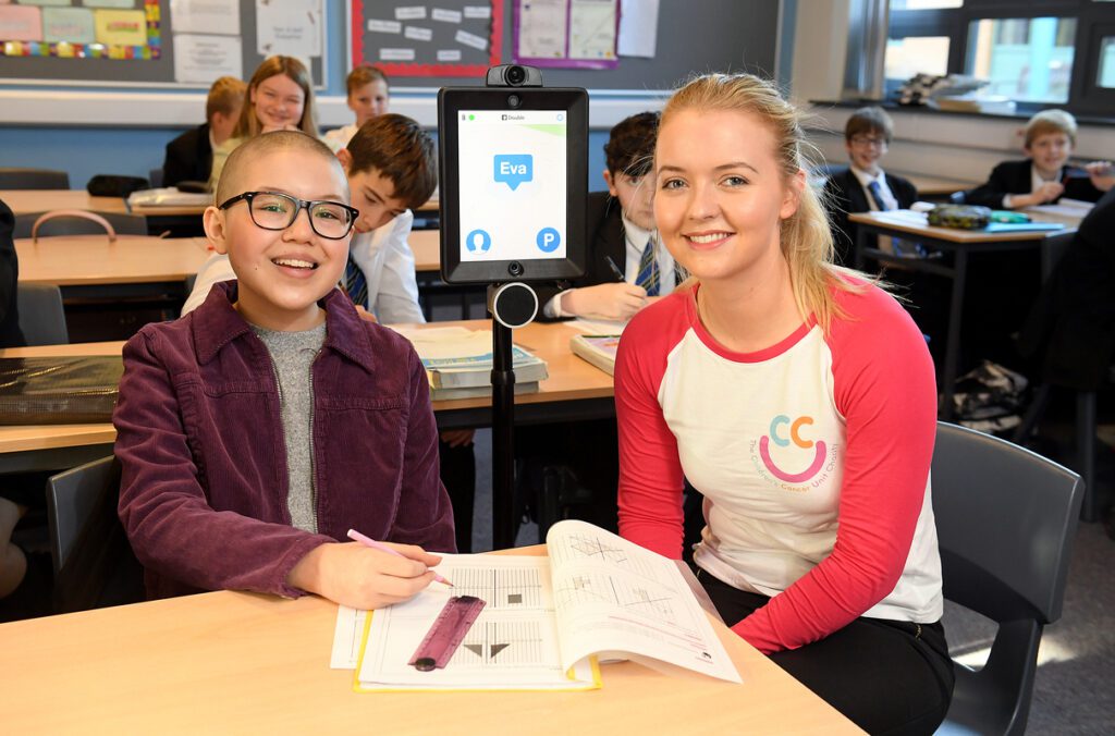 A student sits in a classroom with a tablet on a stand labeled "Eva," joined by a person beside them. Publications and other materials are scattered around, as other students focus intently at their desks in the background.