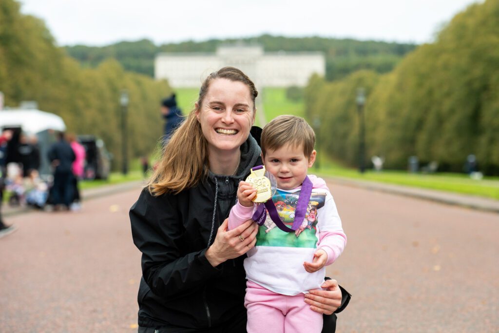 A woman and a child smiling, with the child holding a medal. They are outdoors on a pathway with a large building and trees in the background.