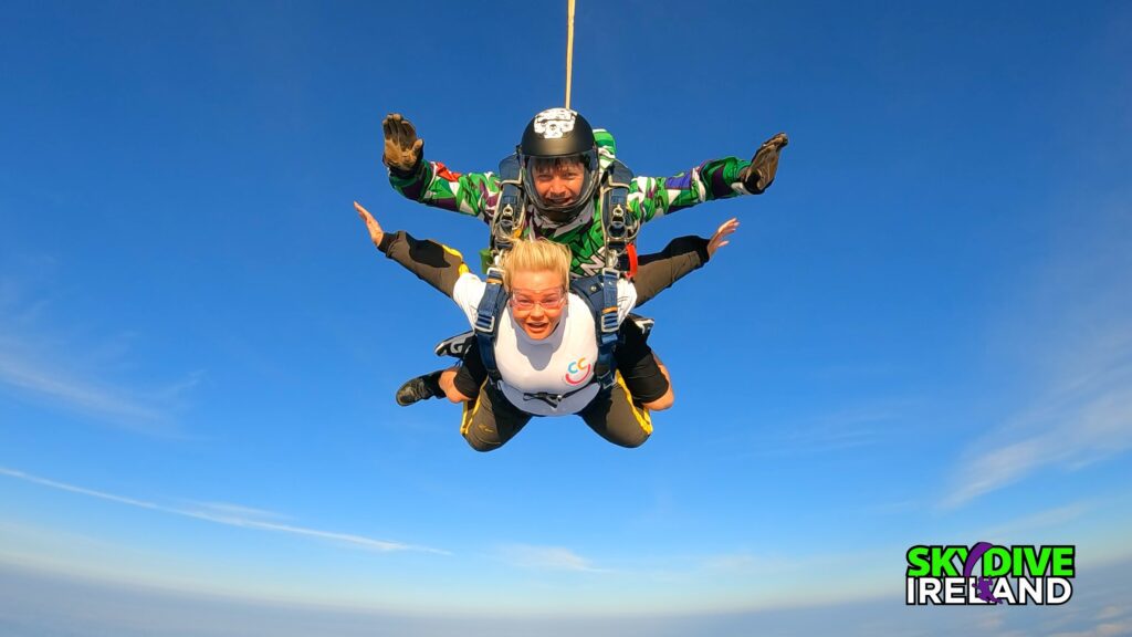Two people tandem skydiving against a clear blue sky, with "Skydive Ireland" logo visible in the bottom right corner.