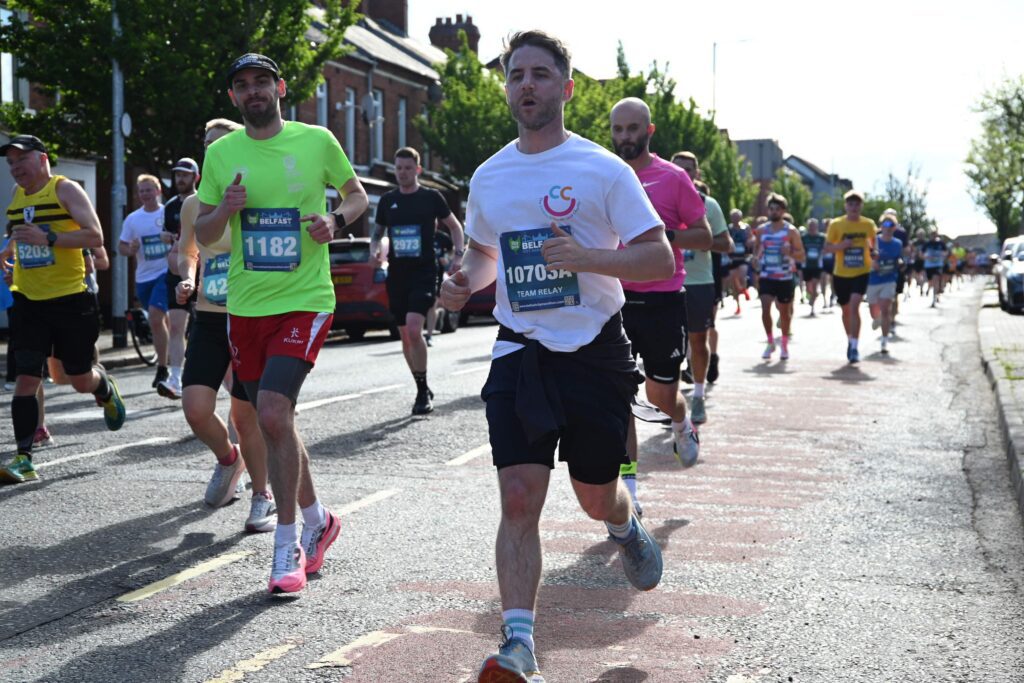 Runners participate in the Belfast City Marathon 2026, wearing numbered bibs and athletic clothing, as they race down streets lined with trees and buildings.