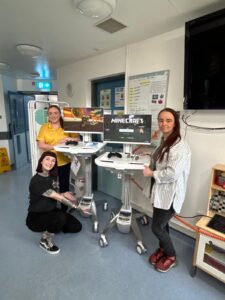 In a supportive gesture to the Children’s Cancer Unit, three people proudly pose with two computers in a room, each displaying a Minecraft screen. Their smiles reflect the joy of gaming and creativity, showcasing the impact of their generous donation.
