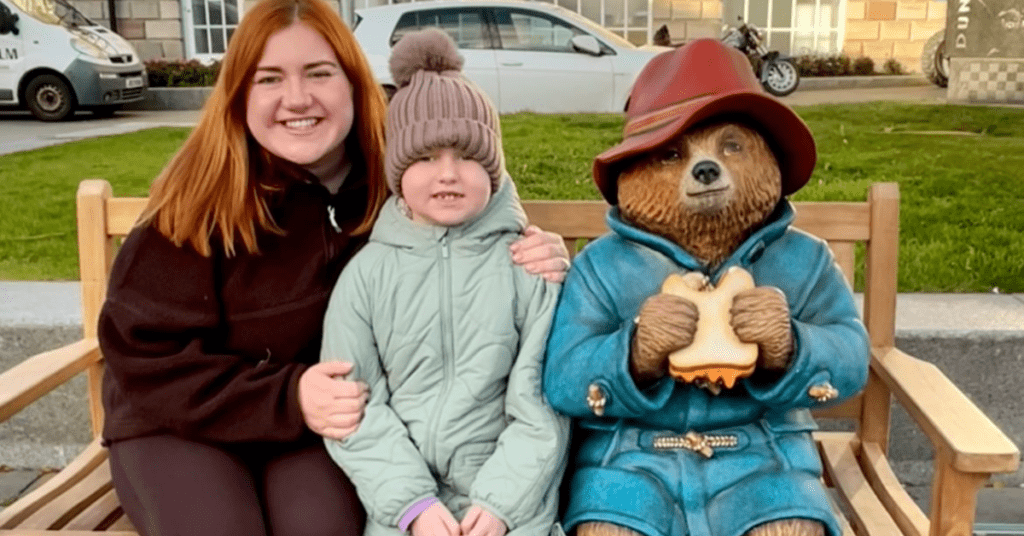 Danielle Smyth and a friend enjoy a quiet moment on a bench beside the iconic Paddington Bear statue, which holds a sandwich as if offering it to passersby. A vehicle hums in the background, adding to this picturesque scene that would make for a perfect Mother’s Day feature.