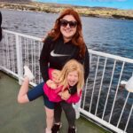 A woman in sunglasses holds a smiling child on a ferry. The background features water and a distant shoreline under a partly cloudy sky.