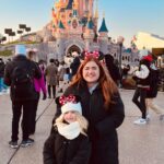 A woman and a child, both wearing Minnie Mouse ears and black coats, stand in front of a castle at a theme park.