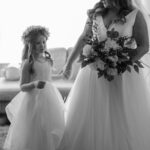 A bride holds a bouquet while standing with a flower girl, both in white dresses, in a softly lit room.