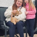 A woman sitting on a chair holding a puppy, with a young girl standing beside her. Both are smiling outdoors against a white textured wall.
