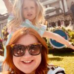 A woman with sunglasses takes a selfie with a young girl posing energetically in front of a large building on a sunny day.