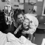 A woman and child smile beside a hospital bed. The woman holds an IV pole. The child is making a playful expression.