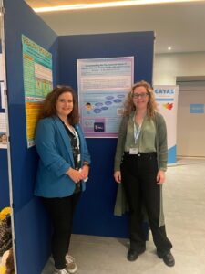 Two women stand in front of a research poster on psychosocial needs at an academic conference, both wearing badges and business-casual clothing.