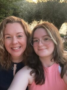 Two people are smiling and posing for a selfie outdoors in natural light, with trees visible in the background, capturing a joyful moment that highlights the importance of meeting psychosocial needs for adolescents and young adults with cancer.