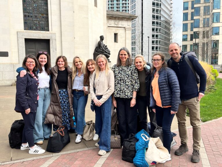 A group of people, part of the CHU team, stand together outdoors, smiling at the camera. They are dressed in casual clothing with bags on the ground nearby. Buildings and a statue are visible in the background, suggesting they might be attending the CCLG Conference for continuous learning.