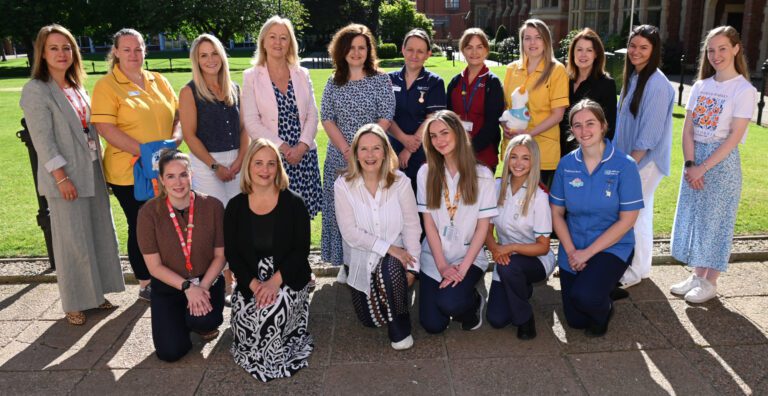 A group of sixteen women, some in professional uniforms, pose for a group photo outdoors on a sunny day at the QUB Student Conference focused on Children’s Cancer.