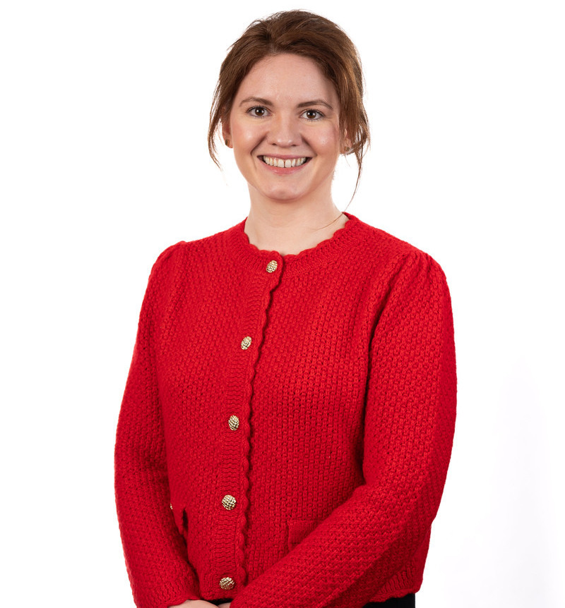 Woman standing and smiling at the camera, wearing a red knit cardigan with gold buttons, in front of a plain white background.