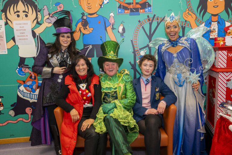 Three characters from a pantomime including a villain, fairy and green man with a top hat sit on a sofa with a mum and teenage daughter.