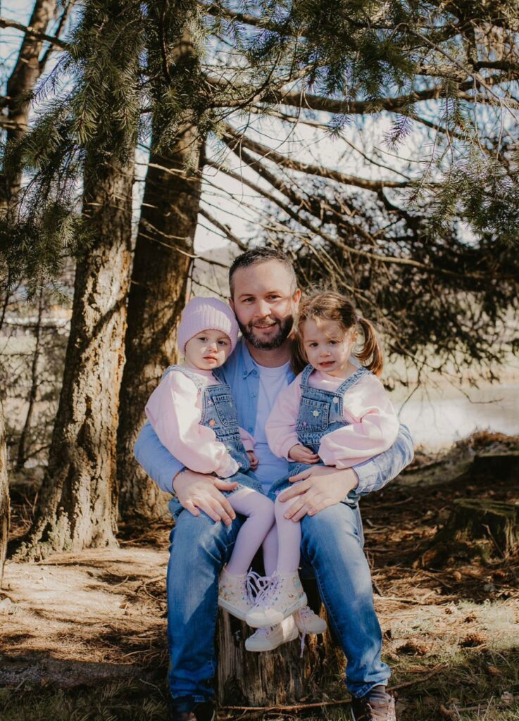 A man sits on a tree stump outdoors with two young girls on his lap, all wearing light blue and pink outfits, surrounded by trees and sunlight—capturing a heartfelt Father’s Day moment from Myla’s Journey.