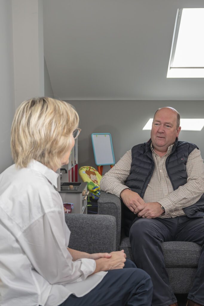 Two people sit facing each other in a room, engaged in conversation. One person has short, light hair and the other wears a dark vest. Children's toys are visible in the background, suggesting a setting for loss support or bereavement support.