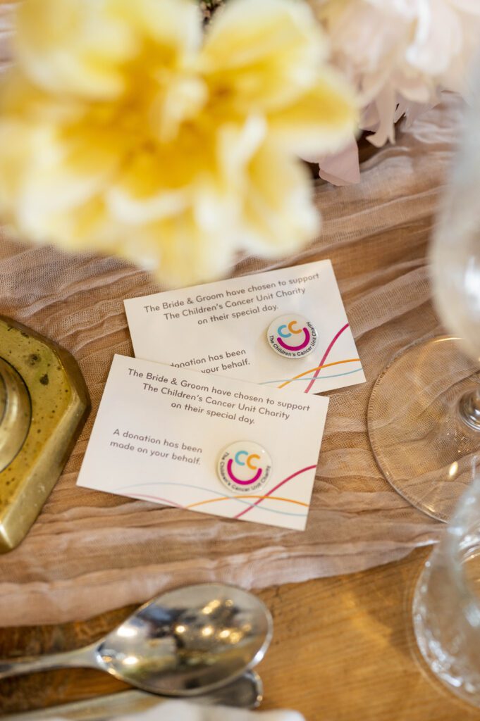 Two Wedding Favours cards displayed on a decorated table announce that a donation has been made to The Children’s Cancer Unit Charity in honour of the wedding guests. Arrangements of flowers, glassware, and cutlery are visible nearby.
